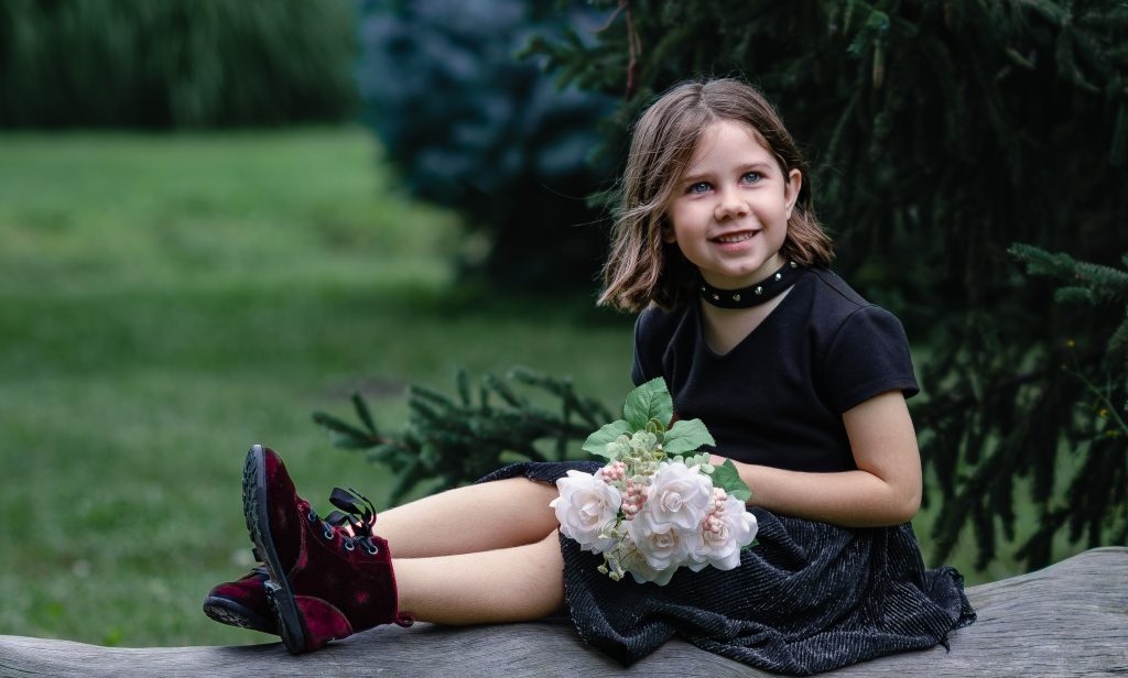 Portrait of a girl sitting on a tree wearing black clothes and red boots holding a bouquet of pink flowers