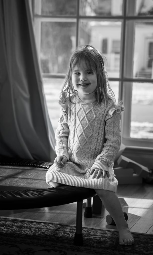 Black and white lifestyle portrait of a little girl smiling wearing a grey dress sitting on a trampoline with a large window behind her. Lyssa Benavidez Photography Elkhorn, NE Lyssab.com lifestyle family photography. 