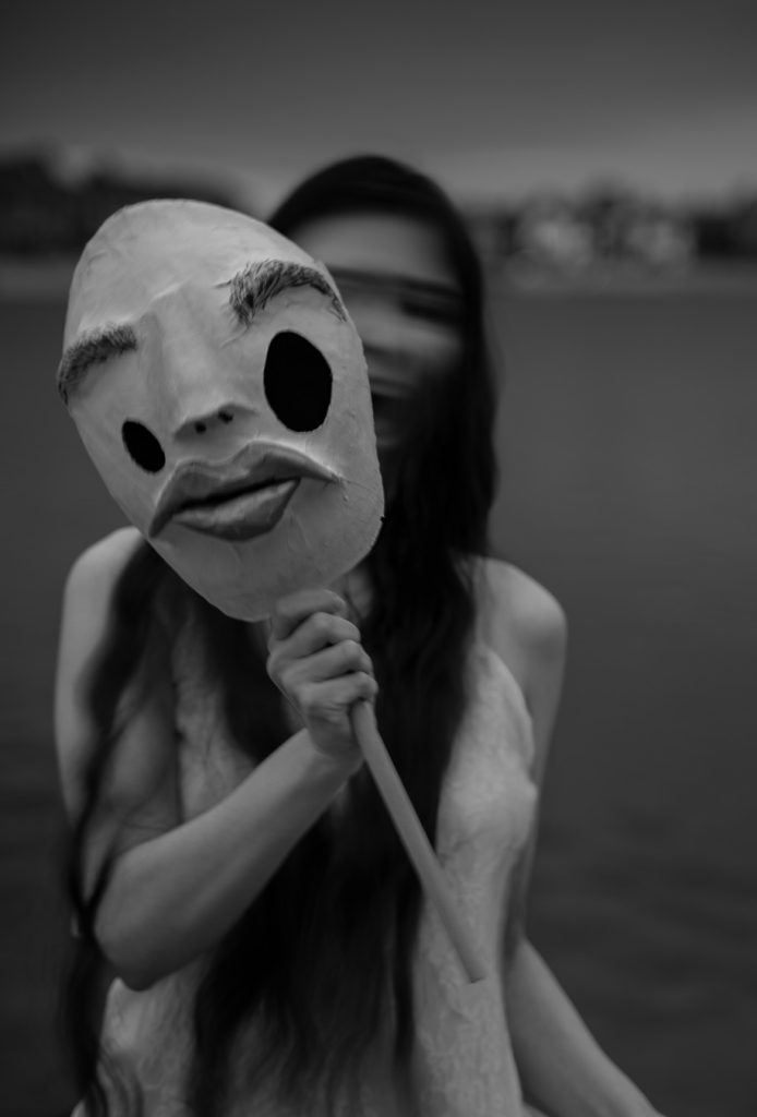 Surrealist black & white portrait of a woman in a white floral dress screaming with motion blur distorting her face holding a creepy humanoid mask in front of her. Lyssa Benavidez Photography Elkhorn, NE Lyssab.com Fine Art Photography Surrealist Photography