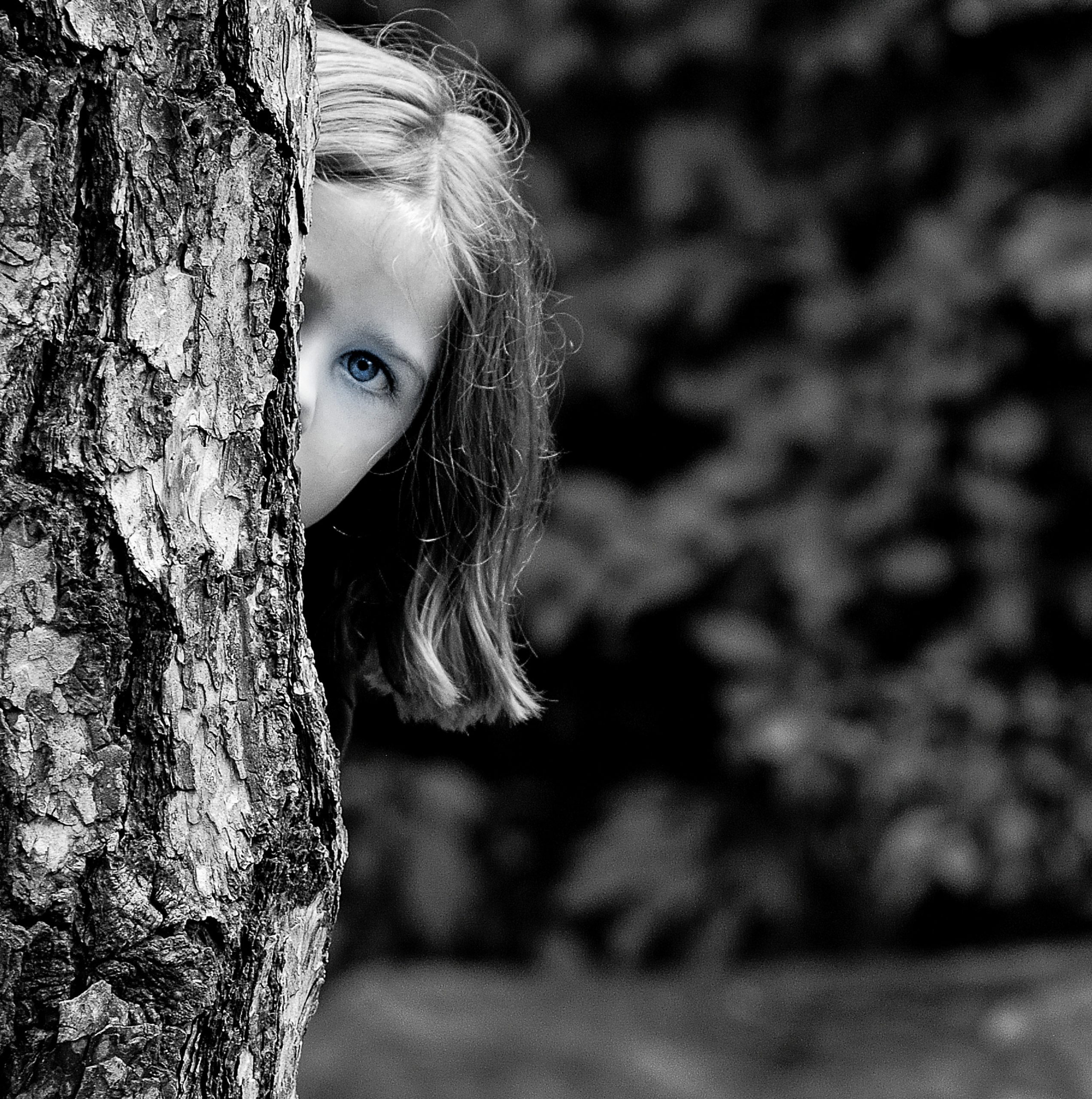 Black & White photo of a little girl looking around a tree with leaves in the background