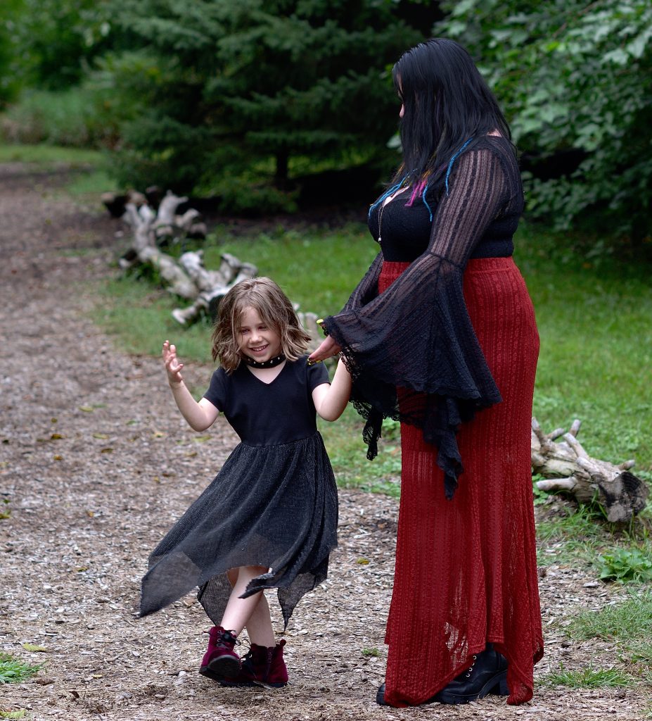 Outdoor family portrait of a mom and her daughter dancing on a tree lined walking path at OPPD abortorium in Omaha NE. The mom is wearing a black long sleeved top with a red skirt and black boots. The girl is wearing a flowy black dress with deep red boots.  Lyssa Benavidez Photography Elkhorn, NE Lyssab.com Family photography. 