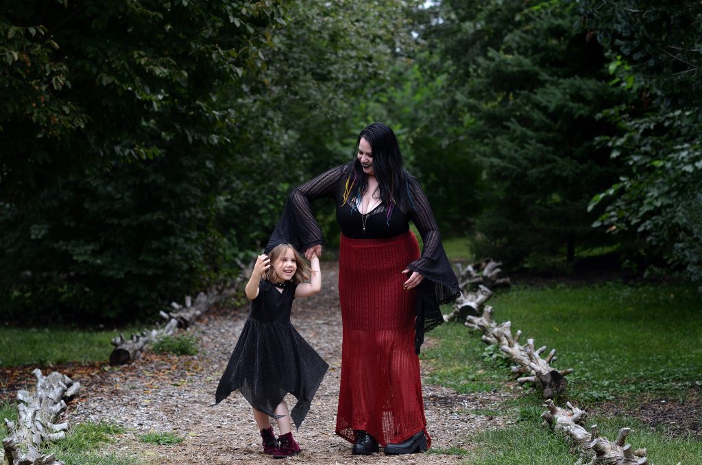Outdoor family portrait of a mom and her daughter dancing on a tree lined walking path at OPPD abortorium in Omaha NE. The mom is wearing a black long sleeved top with a red skirt and black boots. The girl is wearing a flowy black dress with deep red boots.  Lyssa Benavidez Photography Elkhorn, NE Lyssab.com Family photography. 