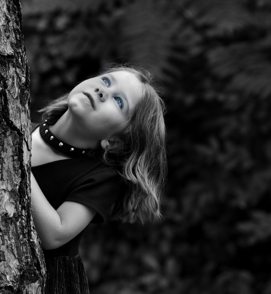 Black & white close up portrait of a girl peeking over a tree trunk with bushes in the background.  Lyssa Benavidez Photography Elkhorn, NE Lyssab.com Family photography. 