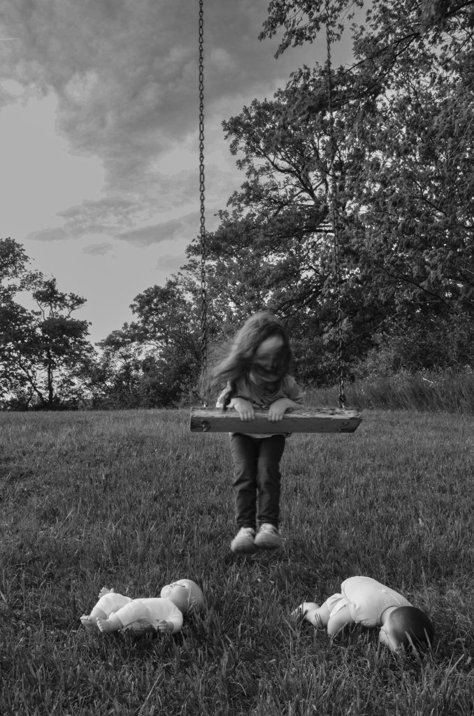 Surrealist portrait of a little girl a t-shirt and dark pants with her hair in her face swinging on an old abandoned swing with two dolls on the grass in front of her and trees and a cloudy sky behind her. Lyssa Benavidez Photography Elkhorn, NE Lyssab.com surrealist photography 