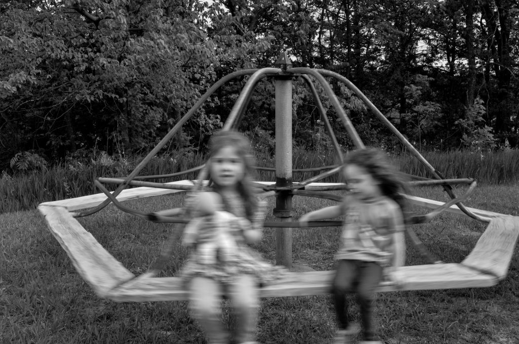 Black and White motion blurred image of two girls on an old merry-go-round with trees in the background