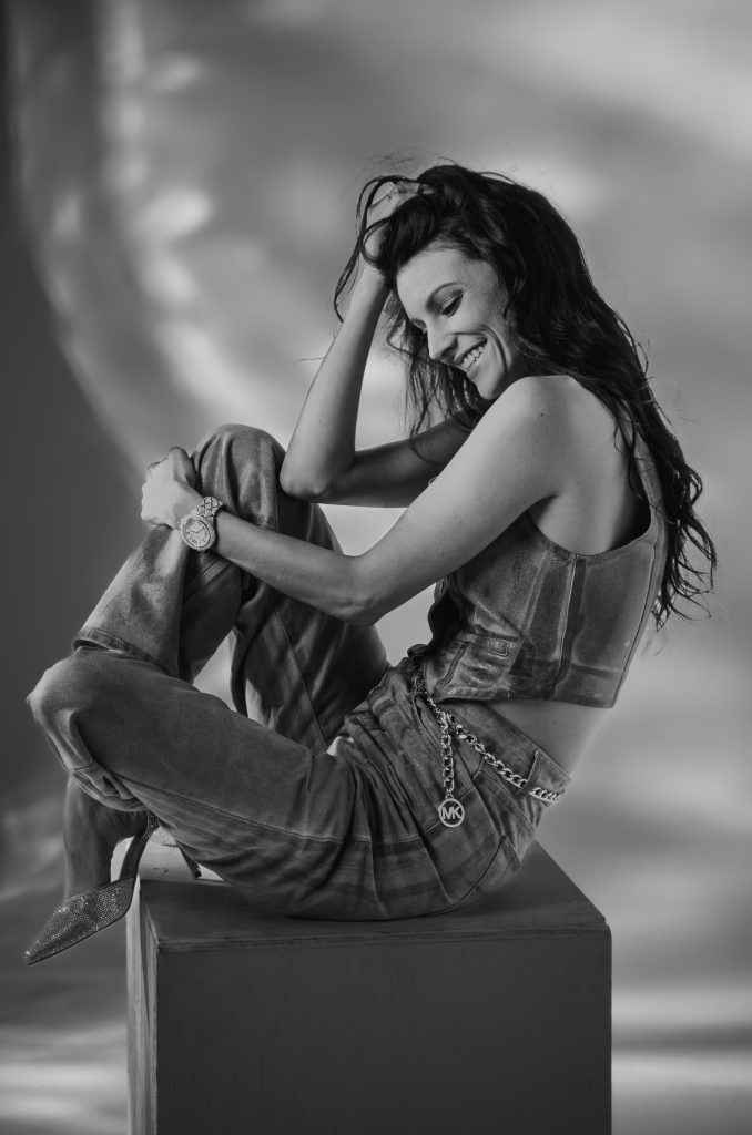 Black and white photo of a woman sitting on a grey box wearing dark clothing with a light and dark lit background. Lyssa Benavidez Photography Elkhorn, NE Lyssab.com studio photography. 