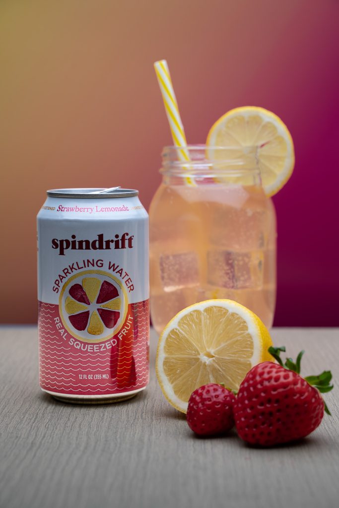 Product photography for Spindrift sparkling water featuring a glass of the pink water with a yellow straw and a lemon slice sticking out of it surrounded by the white and pink can of strawberry lemonade spindrift, half of a yellow lemon and two small red strawberries sitting in front of a yellow and pink background on top of a grey table.  Lyssa Benavidez Photography Elkhorn, NE Lyssab.com Product photography.