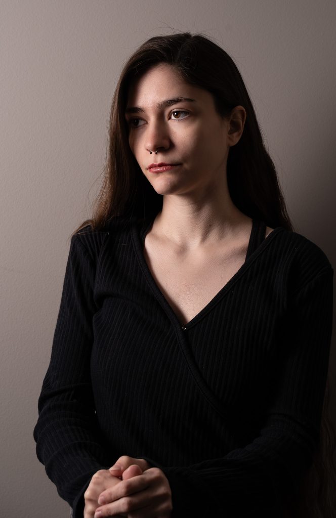 Dramatic portrait of a woman with long hair wearing a black shirt looking off into the distance standing in front of a grey background with dramatic lighting. Lyssa Benavidez Photography Elkhorn, NE Lyssab.com studio fine art photography