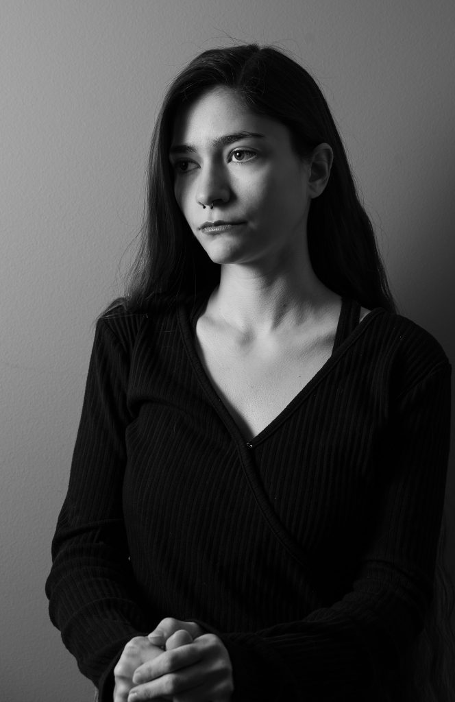 Black and white portrait of a woman with long hair wearing a black shirt looking off into the distance standing in front of a grey background with dramatic lighting. Lyssa Benavidez Photography Elkhorn, NE Lyssab.com studio fine art photography