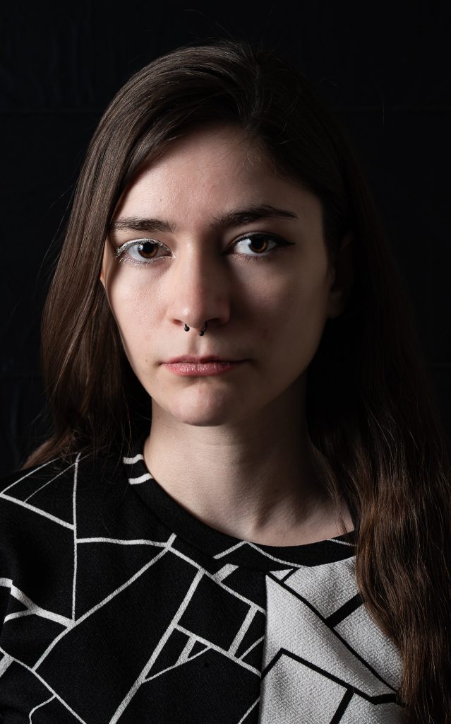 Dramatic portrait of a woman with long hair wearing a black and white shirt looking at the camera standing in front of a black background with dramatic lighting. Lyssa Benavidez Photography Elkhorn, NE Lyssab.com studio fine art photography