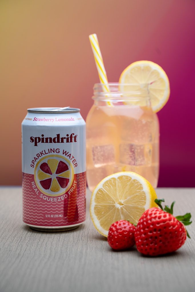 Product photography for Spindrift sparkling water featuring a glass of the pink water with a yellow straw and a lemon slice sticking out of it surrounded by the white and pink can of strawberry lemonade spindrift, half of a yellow lemon and two small red strawberries sitting in front of a yellow and pink background on top of a grey table.  Lyssa Benavidez Photography Elkhorn, NE Lyssab.com Product photography.
