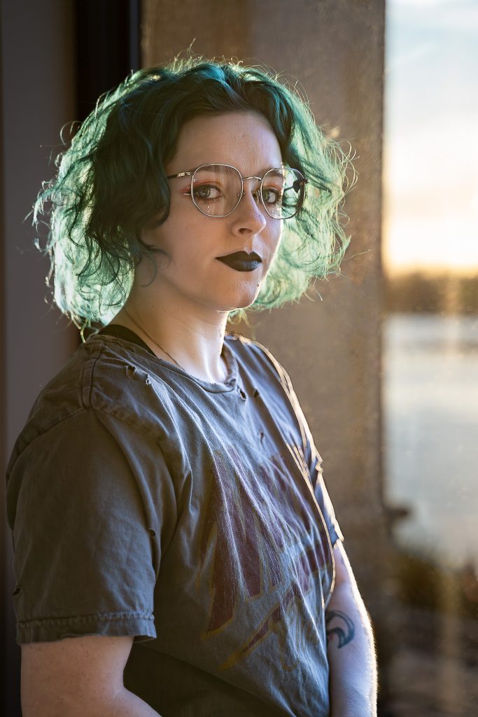 portrait of a woman with blue/green hair wearing glasses and dark lipstick, a grey shirt standing in front of a window with a body of water and a warm sunset in the background. Lyssa Benavidez Photography Elkhorn, NE Lyssab.com Self-portrait photography