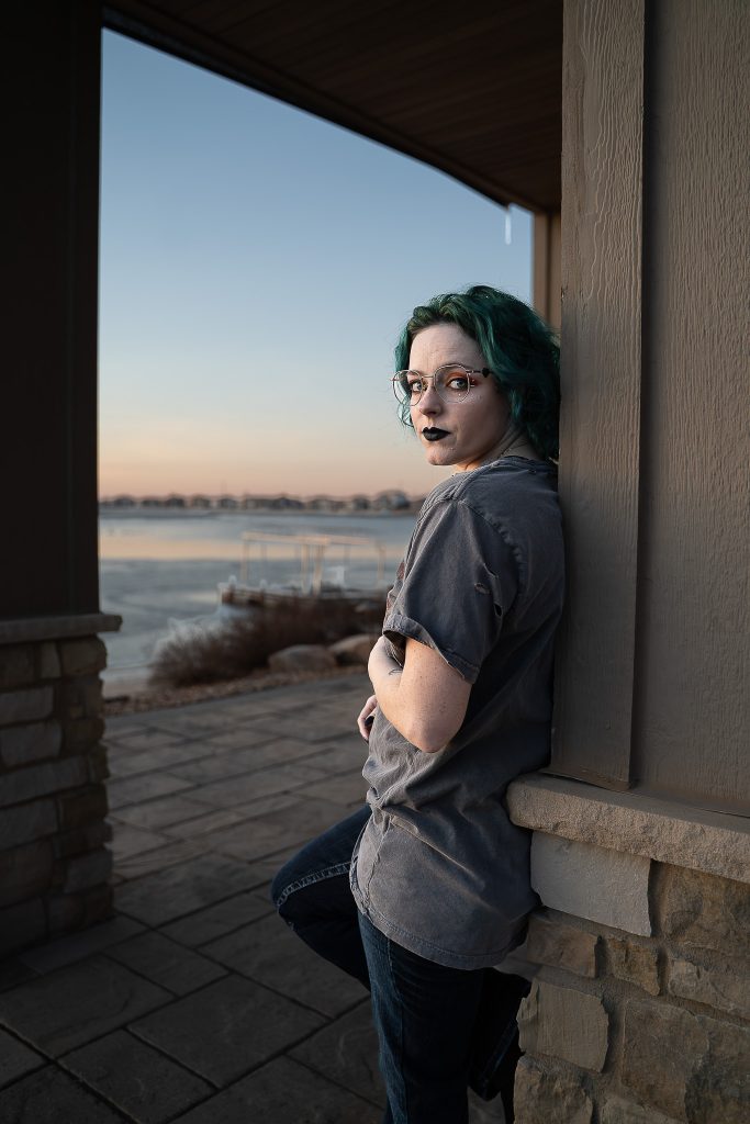 Portrait of a woman wearing glasses and dark lipstick, a grey shirt, and dark pants leaning against a wall with a body of water in the background. Lyssa Benavidez Photography Elkhorn, NE Lyssab.com Self-portrait photography