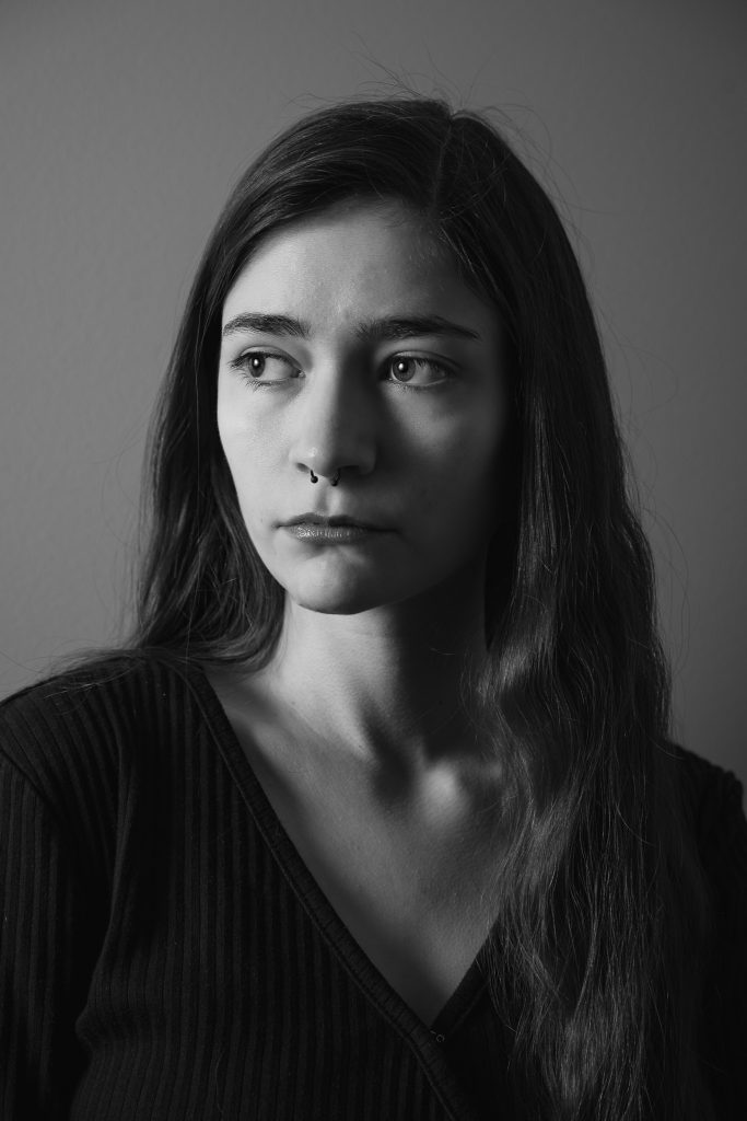 Black and white portrait of a woman with long hair wearing a black shirt looking off into the distance standing in front of a grey background with dramatic lighting. Lyssa Benavidez Photography Elkhorn, NE Lyssab.com studio fine art photography