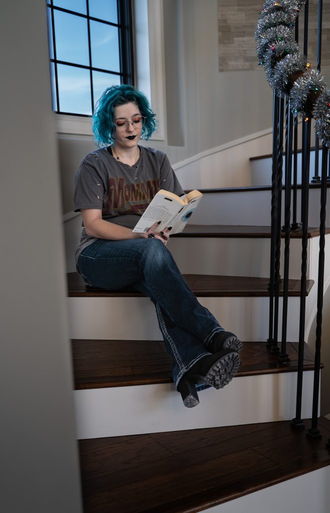 portrait of a woman with blue hair wearing glasses and dark lipstick, a grey shirt, dark pants, and black boots sitting on brown and white stairs reading a white and blue book with a window behind her. Lyssa Benavidez Photography Elkhorn, NE Lyssab.com Self-portrait photography