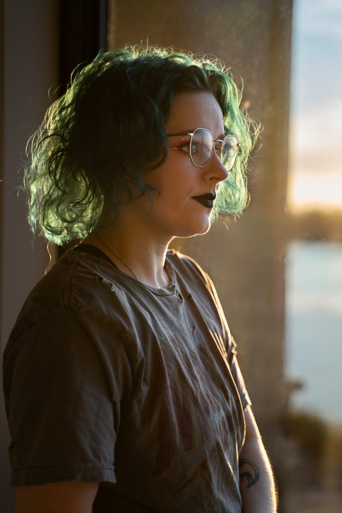 portrait of a woman with blue/green hair wearing glasses and dark lipstick, a grey shirt standing in front of a window with a body of water and a warm sunset in the background staring off into the distance. Lyssa Benavidez Photography Elkhorn, NE Lyssab.com Self-portrait photography