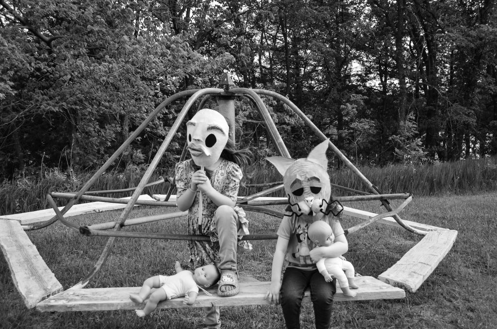 Ralph Eugune Meatyard style creative black and white portrait of two little kids wearing eerie humanoid facemasks sitting on an old abandoned merry-go-round with trees in the background in Plattsmouth NE. Lyssa Benavidez Photography Elkhorn, NE Lyssab.com Creative/Fine art photography. 