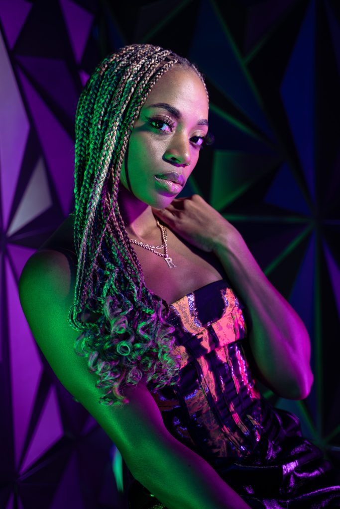 Creative light photo of a woman sitting on a stool wearing dark colored, shiny clothing with pink and green light and a geometric green and purple background. Lyssa Benavidez Photography Elkhorn, NE Lyssab.com studio photography. 
