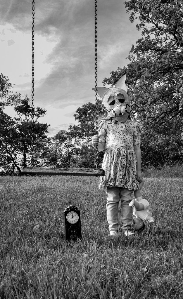 Surrealist portrait of a little girl wearing a floral dress and light colored pants wearing a creepy humanoid mask holding an old abandoned swing in one hand and a doll in the other. There is a small clock on the grass under the swing and trees and a cloudy sky behind her. Lyssa Benavidez Photography Elkhorn, NE Lyssab.com surrealist photography 