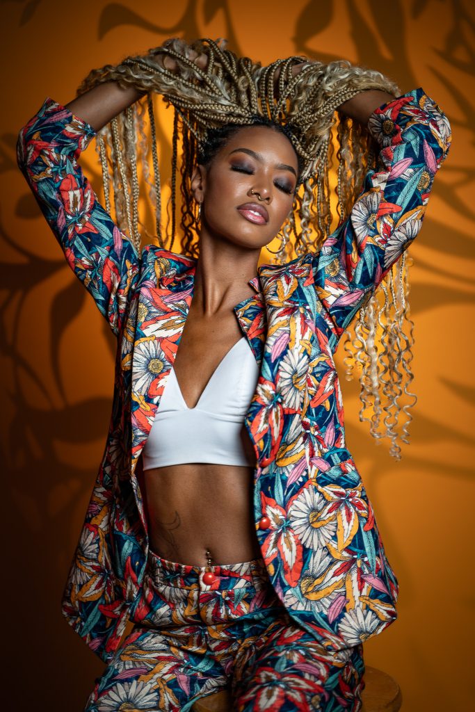 Creative studio portrait of a woman holding her braided hair above her hair with her eyes closed wearing a colorful floral outfit with a cropped white top underneath standing in front of a dark yellow background. Lyssa Benavidez Photography Elkhorn, NE Lyssab.com studio photography. 