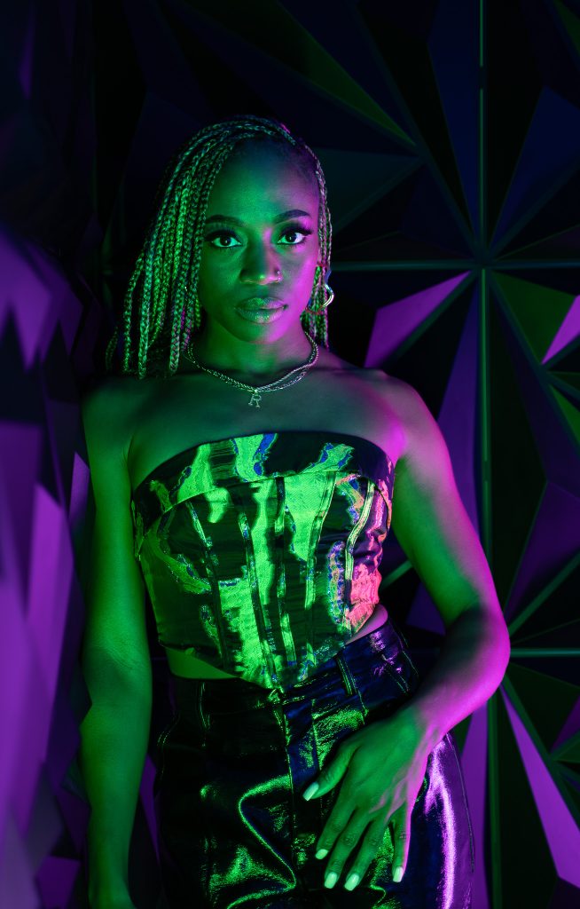 Creative light photo of a woman leaning against a wall wearing dark colored, shiny clothing with pink and green light and a geometric green and purple background. Lyssa Benavidez Photography Elkhorn, NE Lyssab.com studio photography. 