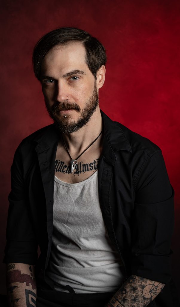 Portrait of a man with a menacing look wearing a black and white shirt with dramatic studio lighting standing in front of a red background. Lyssa Benavidez Photography Elkhorn, NE studio photography. 