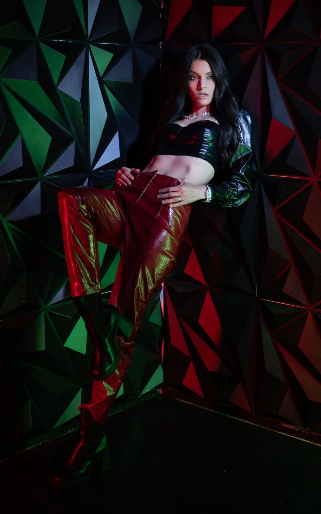 Creative light photo of a woman leaning against a wall wearing red and black colored, shiny clothing with red and green light and a geometric green and red background. Lyssa Benavidez Photography Elkhorn, NE Lyssab.com studio photography. 