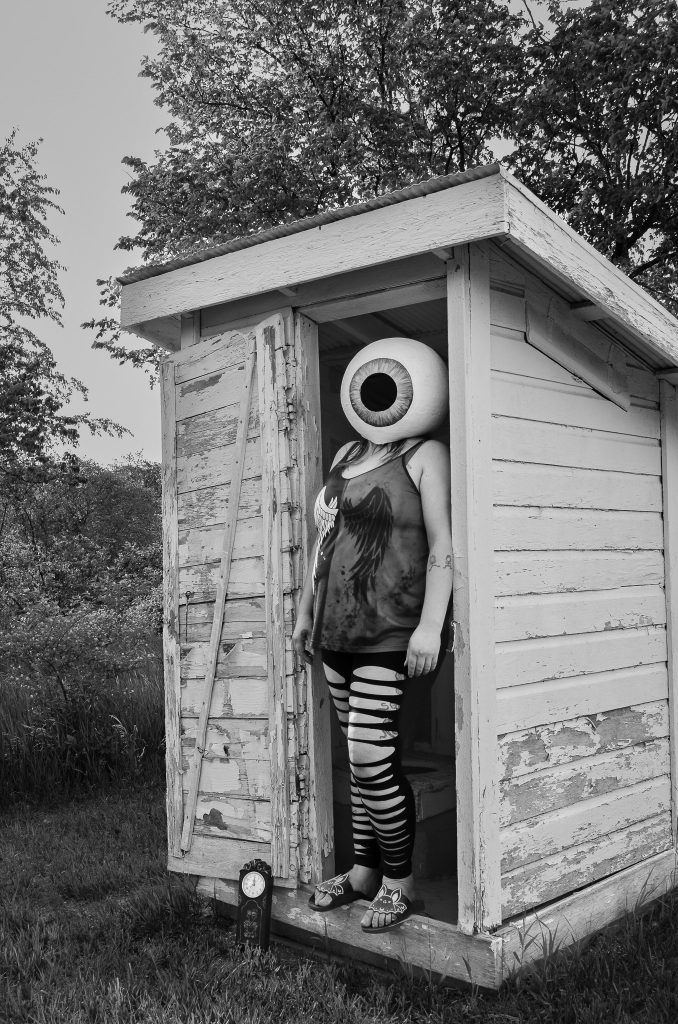 Surrealist portrait of a woman standing in an abandoned outhouse wearing a tank top and ripped black pants wearing an eyeball mask on her head with a clock at her feet and trees and a sky in the background. Lyssa Benavidez Photography Elkhorn, NE Lyssab.com surrealist photography 