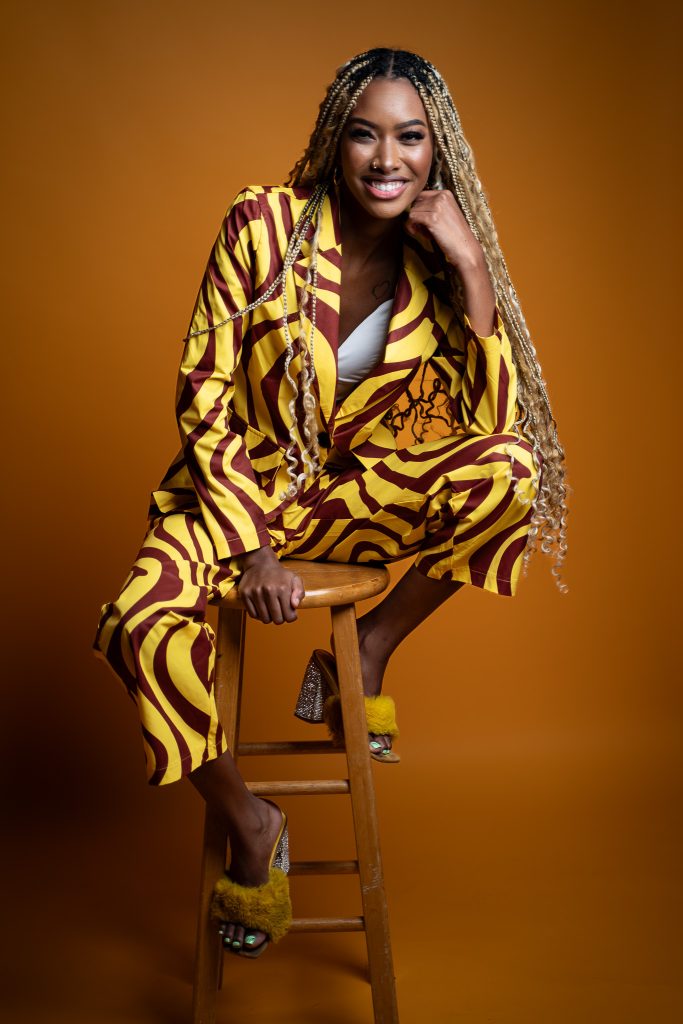 Creative studio portrait of a woman sitting on a stool wearing a yellow and brown outfit with a cropped white top underneath standing in front of a dark yellow background. Lyssa Benavidez Photography Elkhorn, NE Lyssab.com studio photography. 