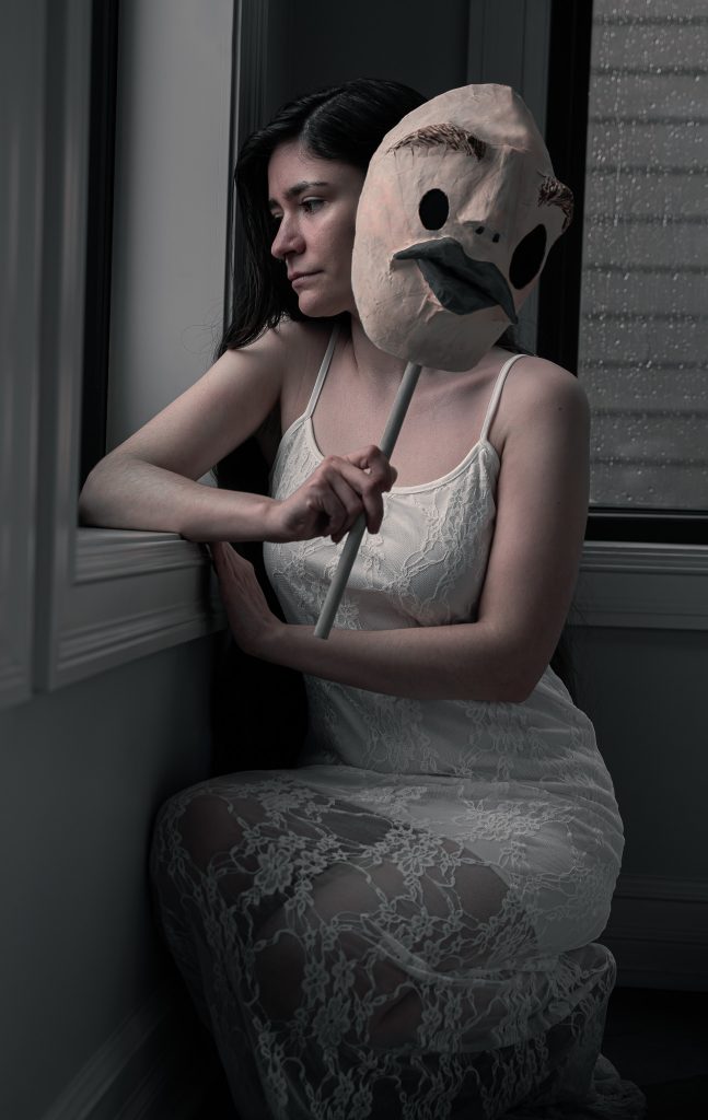 Surrealist fine art photo of a woman in a white floral dress somberly looking out the window holding a creepy humanoid mask.  Lyssa Benavidez Photography Elkhorn, NE Lyssab.com Fine Art Photography surrealist photography