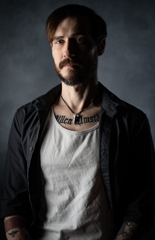 Dramatic studio portrait of a man with a curious look wearing a white and black shirt with dramatic studio lighting standing in front of a grey background. Lyssa Benavidez Photography Elkhorn, NE studio photography. 