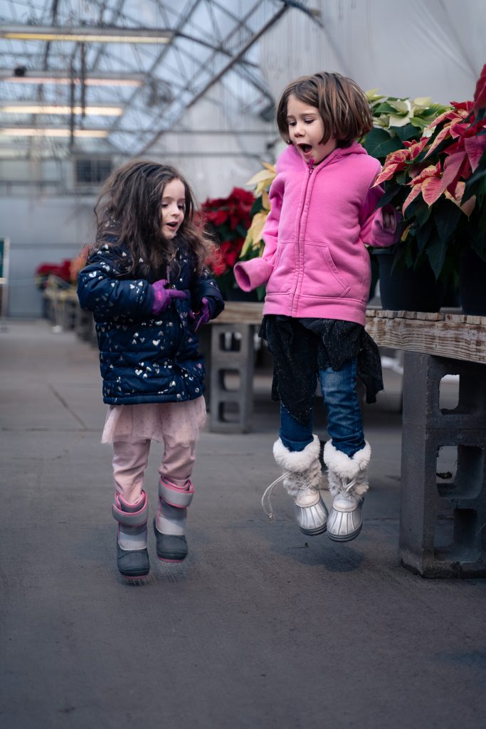 Portrait of two girls jumping in an aisle together causing intentional motion blur with plants in the background. Lyssa Benavidez Photography Elkhorn, NE Lyssab.com Lifestyle family photography