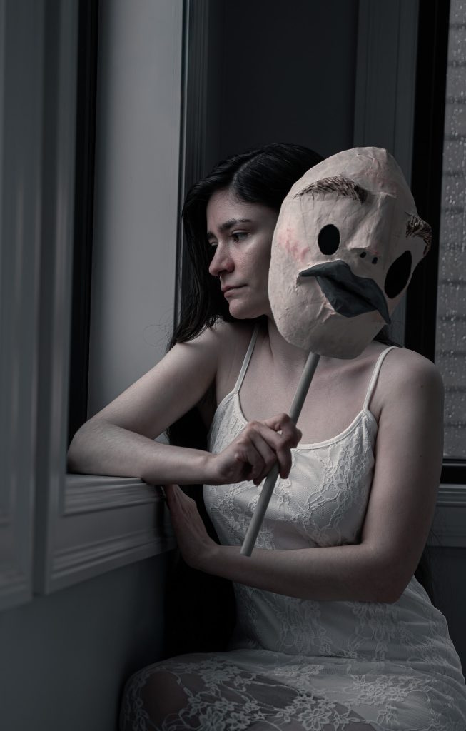 Surrealist fine art photo of a woman in a white floral dress somberly looking out the window holding a creepy humanoid mask.  Lyssa Benavidez Photography Elkhorn, NE Lyssab.com Fine Art Photography surrealist photography