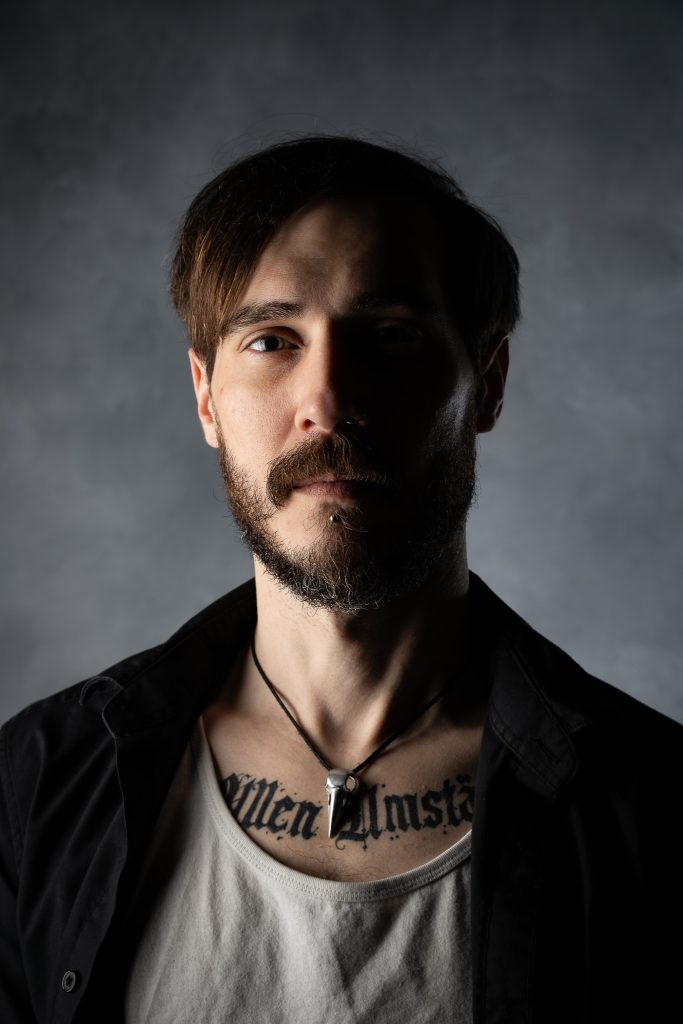 Dramatic portrait of a man with a somber look wearing a white and black shirt with dramatic studio lighting standing in front of a grey background. Lyssa Benavidez Photography Elkhorn, NE studio photography. 