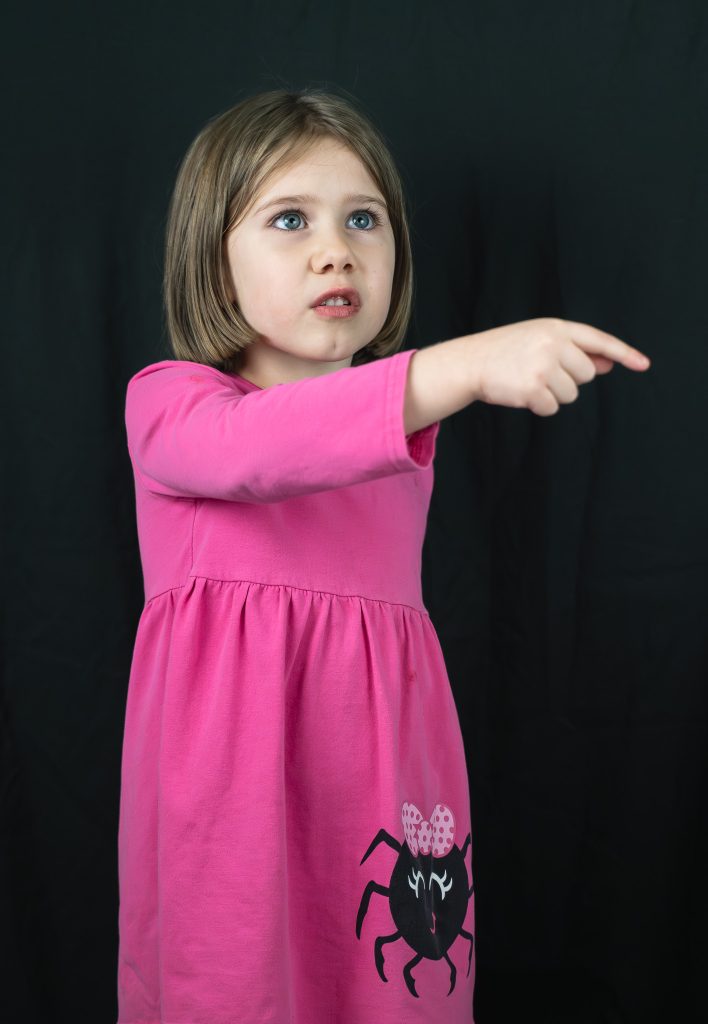 Studio portrait of a little girl with brown hair pointing to something off camera looking worried wearing a pink dress with a black spider on the front right side standing in front of a black background. Lyssa Benavidez Photography Elkhorn, NE Lyssab.com Studio photography. 