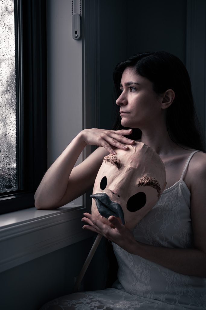 Surrealist fine art photo of a woman in a white floral dress somberly looking out the window holding a creepy humanoid mask.  Lyssa Benavidez Photography Elkhorn, NE Lyssab.com Fine Art Photography surrealist photography