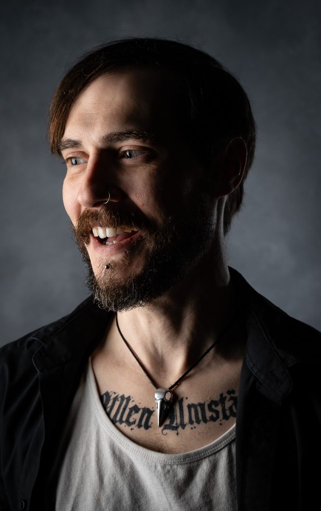 Photo of a man with a joyful expression wearing a black and white shirt with dramatic lighting standing in front of a grey background. Lyssa Benavidez Photography Elkhorn, NE  