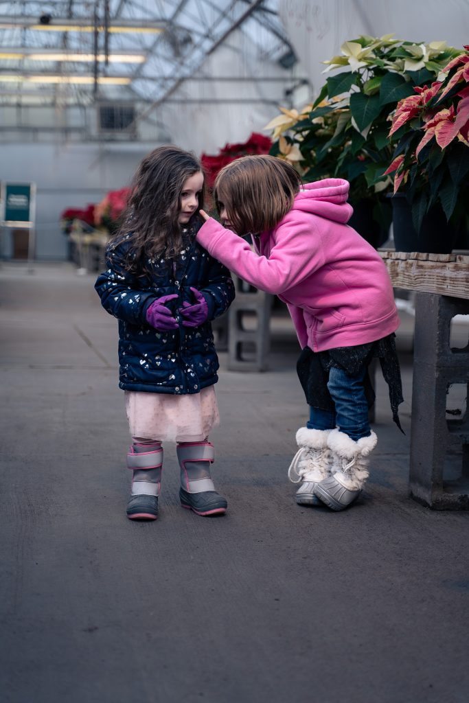 Portrait of two girls talking in an aisle together intensely with plants in the background. Lyssa Benavidez Photography Elkhorn, NE Lyssab.com Lifestyle family photography