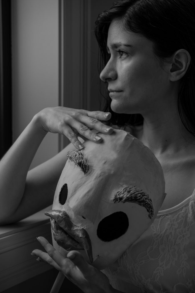 Surrealist fine art black & white photo of a woman in a white floral dress somberly looking out the window holding a creepy humanoid mask.  Lyssa Benavidez Photography Elkhorn, NE Lyssab.com Fine Art Photography surrealist photography