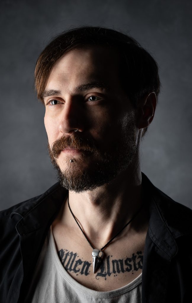Dramatic portrait of a man with a somber look wearing a white and black shirt with dramatic studio lighting standing in front of a grey background. Lyssa Benavidez Photography Elkhorn, NE studio photography. 