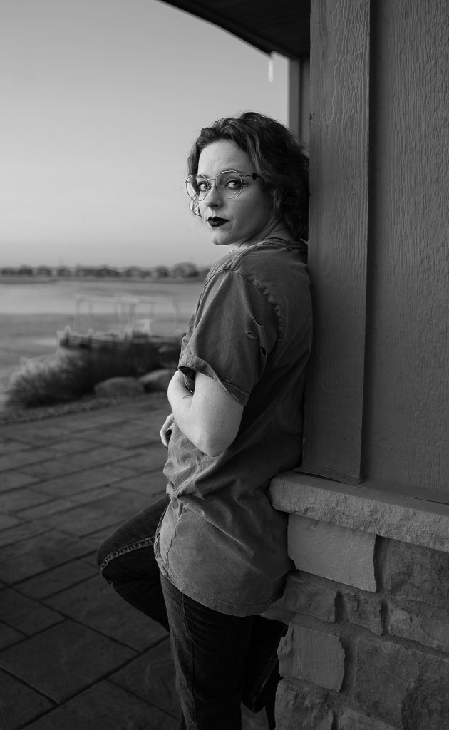 Black and white portrait of a woman wearing glasses and dark lipstick, a grey shirt, and dark pants leaning against a wall with a body of water in the background. Lyssa Benavidez Photography Elkhorn, NE Lyssab.com Self-portrait photography