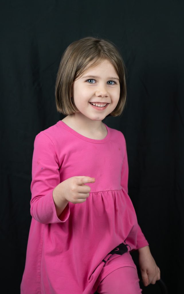 Studio portrait of a little girl with brown hair wearing a pink dress and pants pointing at the camera and smiling. Lyssa Benavidez Photography Elkhorn, NE Lyssab.com studio photography