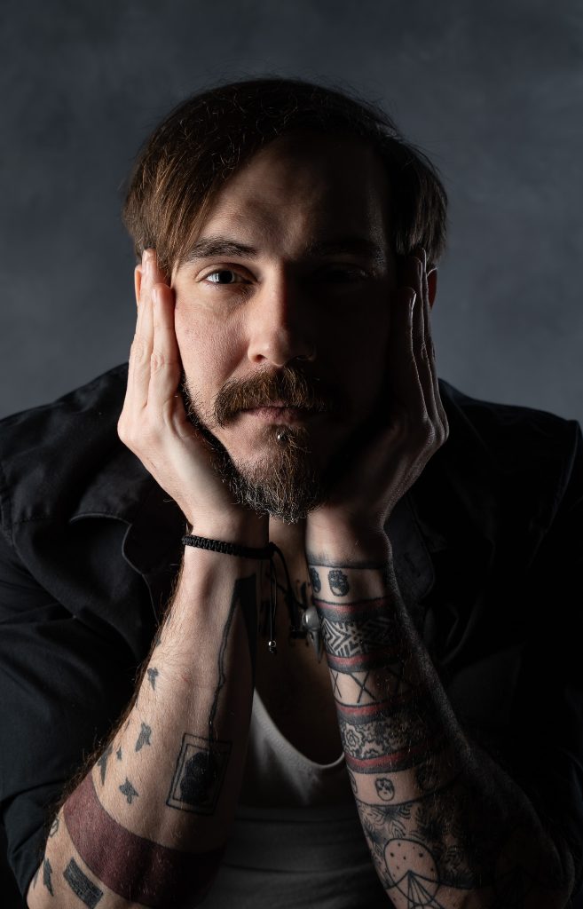 Dramatic studio portrait of a man with a curious look wearing a white and black shirt with dramatic studio lighting standing in front of a grey background. Lyssa Benavidez Photography Elkhorn, NE studio photography. 