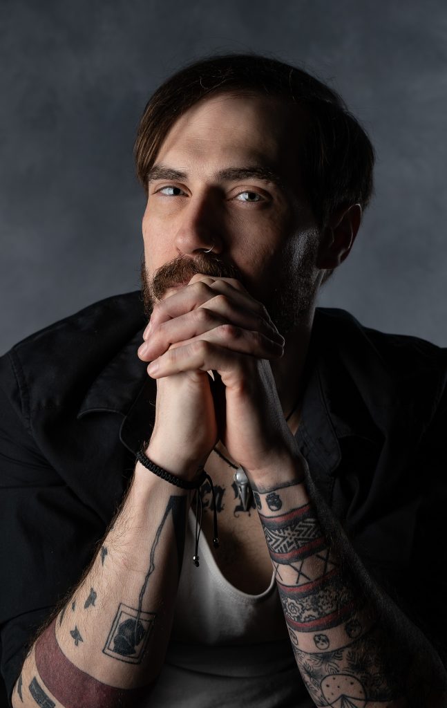 Dramatic studio portrait of a man with a curious look wearing a black and white shirt with tattoos standing in front of a grey background. Lyssa Benavidez Photography Elkhorn, NE studio photography. Lyssab.com
