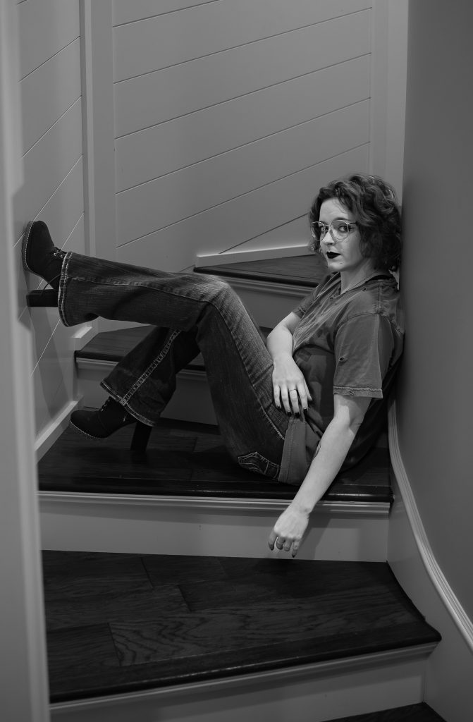 Black and white portrait of a woman wearing glasses and dark lipstick, a grey shirt, and dark pants sitting on white and brown stairs with a white wall behind her. Lyssa Benavidez Photography Elkhorn, NE Lyssab.com Self-portrait photography