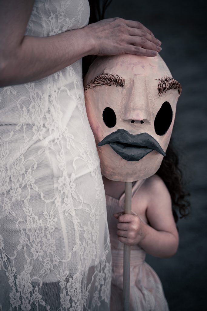 Surrealist portrait of a woman in a white floral dress standing next to a little girl in a pink and white striped outfit. The little girl is holding a creepy humanoid mask.  Lyssa Benavidez Photography Elkhorn, NE Lyssab.com Surrealist Photography   