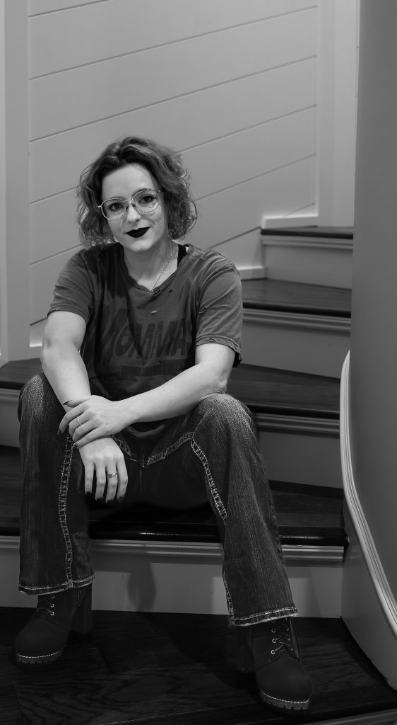Black and white portrait of a woman wearing glasses and dark lipstick, a grey shirt, dark pants, and black boots sitting on brown and white stairs staring into the camera with a white wall behind her. Lyssa Benavidez Photography Elkhorn, NE Lyssab.com Self-portrait photography