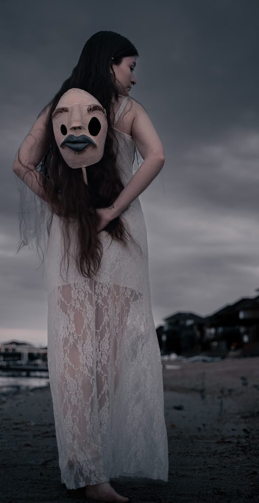 Surrealist portrait of a woman standing on a beach, her back facing us wearing a white floral dress holding a creepy humanoid mask behind her.  Lyssa Benavidez Photography Elkhorn, NE Lyssab.com Fine Art Photography Surrealist photography