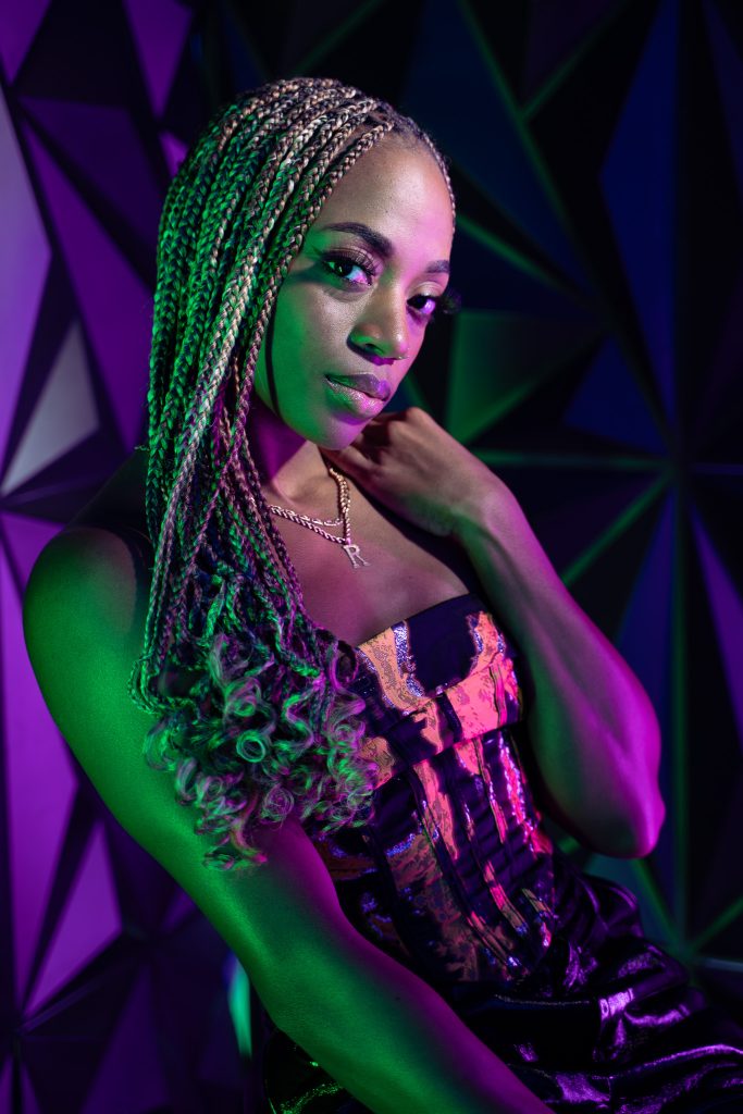 Creative light photo of a woman sitting on a stool wearing dark colored, shiny clothing with pink and green light and a geometric green and purple background. Lyssa Benavidez Photography Elkhorn, NE Lyssab.com studio photography. 