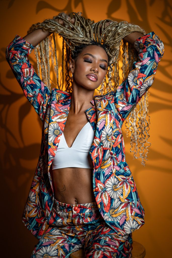 Creative studio portrait of a woman holding her braided hair above her hair with her eyes closed wearing a colorful floral outfit with a cropped white top underneath standing in front of a dark yellow background. Lyssa Benavidez Photography Elkhorn, NE Lyssab.com studio photography. 
